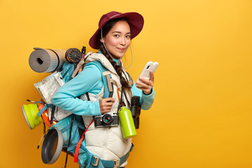 Pretty satisfied traveler uses free internet connection on smartphone for blogging during wanderlust trip, carries big heavy rucksack, has binoculars and retro camera to explore surroundings