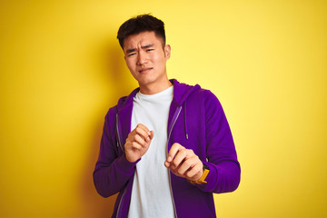 Young asian chinese man wearing purple sweatshirt standing over isolated yellow background disgusted expression, displeased and fearful doing disgust face because aversion reaction. With hands raised