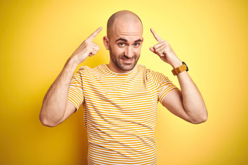 Young bald man with beard wearing casual striped t-shirt over yellow isolated background smiling...