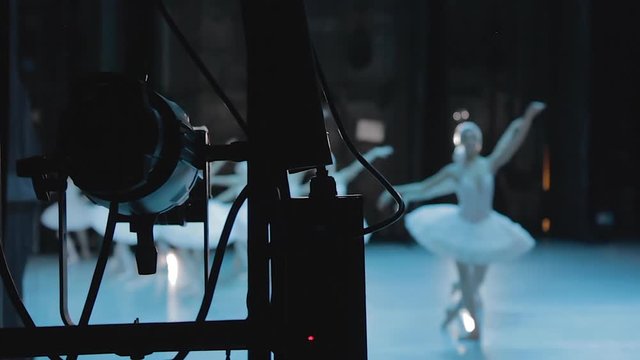Young beautiful ballerinas dance and perform on stage of ballet theater. Dancers wearing pointe shoes and white costumes show classic swan dance in interior with blue light. Video shooting is in room