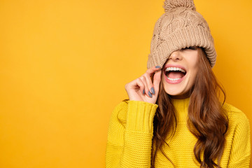 A brunette stands on a yellow background in a yellow sweater, pulls the hat down and laughs joyfully