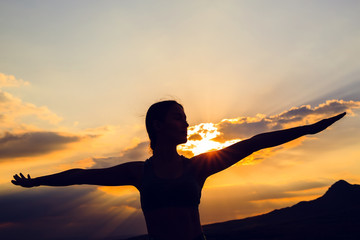 Fototapeta premium Silhouette of young woman practicing yoga or pilates at sunset or sunrise in beautiful mountain location.