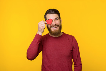 Handsome bearded man, in red sweater fell in love and holding a little heart over eye, like a gesture, standing over yellow background