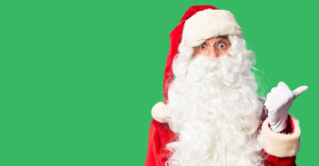 Middle age handsome man wearing Santa Claus costume and beard standing Surprised pointing with hand finger to the side, open mouth amazed expression.