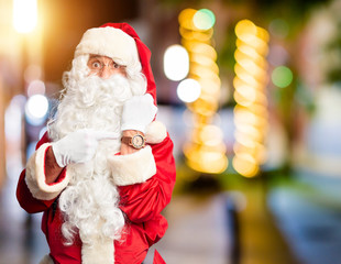Middle age handsome man wearing Santa Claus costume and beard standing In hurry pointing to watch time, impatience, looking at the camera with relaxed expression