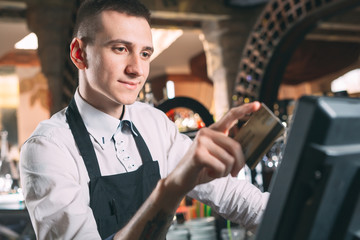 small business, people and service concept - happy man or waiter in apron at counter with cashbox working at bar or coffee shop