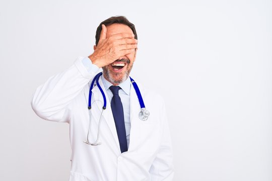 Middle age doctor man wearing coat and stethoscope standing over isolated white background smiling and laughing with hand on face covering eyes for surprise. Blind concept.
