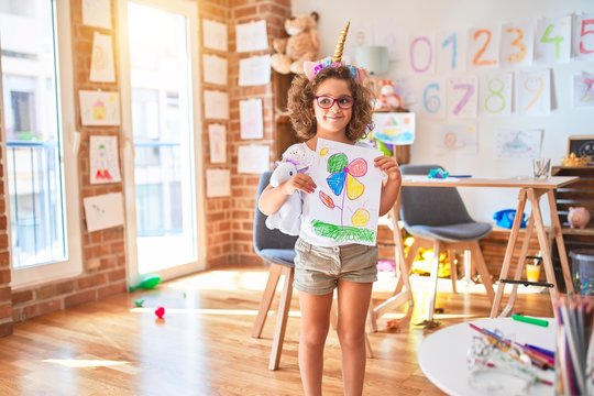 Beautiful toddler wearing glasses and unicorn diadem standing holding draw smiling at kindergarten