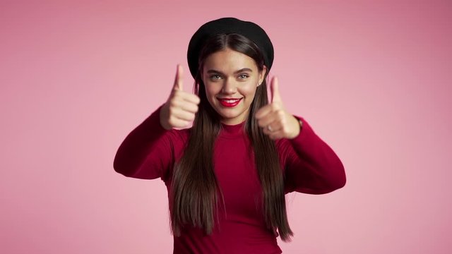 Young hipster woman with red lips and hat making thumbs up sign over pink background. Winner. Success. Positive girl smiles to camera. Body language. Slow motion.