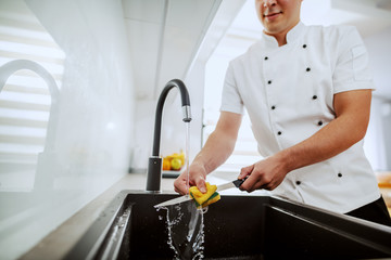 Cropped picture of caucasian chef in uniform washing kitchen knife in sink. Kitchen interior.