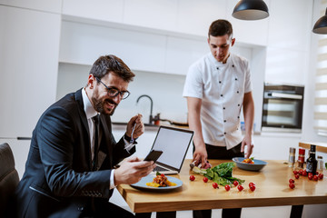 Attractive caucasian bearded businessman in suit using smart phone and eating meal while sitting at dining table. In background is chef serving meals.
