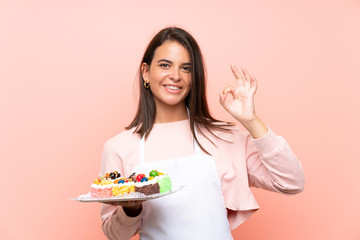 Young girl holding lots of different mini cakes over isolated background showing ok sign with fingers