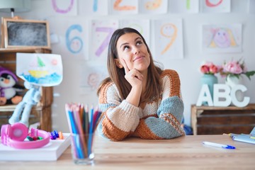 Young beautiful teacher woman wearing sweater and glasses sitting on desk at kindergarten with hand on chin thinking about question, pensive expression. Smiling with thoughtful face. Doubt concept. - obrazy, fototapety, plakaty