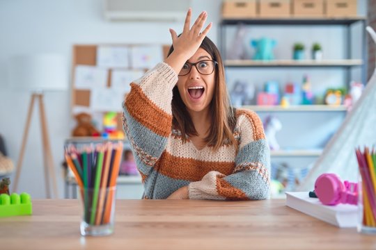 Young beautiful teacher woman wearing sweater and glasses sitting on desk at kindergarten surprised with hand on head for mistake, remember error. Forgot, bad memory concept.