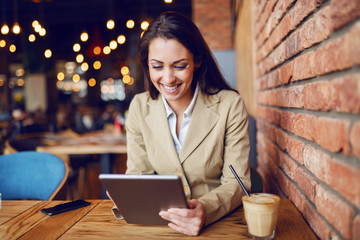 Beautiful caucasian brunette dressed smart casual sitting in cafe and using tablet. On desk are smart phone and coffee.