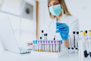 Close up of lab assistant in uniform, with mask and rubber gloves holding test tube with blood sample while sitting on chair and typing on laptop. Selective focus on test tubes. - obrazy, fototapety, plakaty