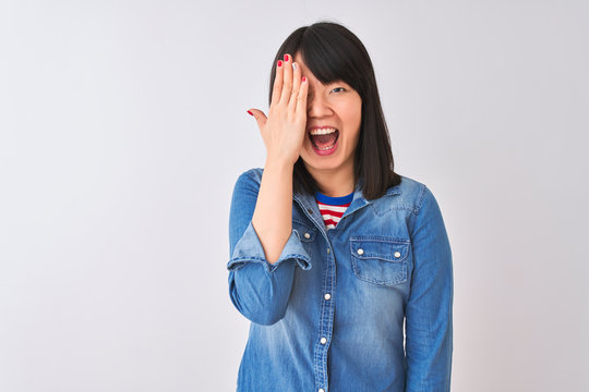 Young beautiful chinese woman wearing denim shirt over isolated white background covering one eye with hand, confident smile on face and surprise emotion.