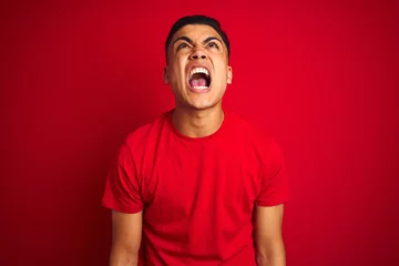 Fotobehang Young brazilian man wearing t-shirt standing over isolated red background angry and mad screaming frustrated and furious, shouting with anger. Rage and aggressive concept. © Krakenimages.com