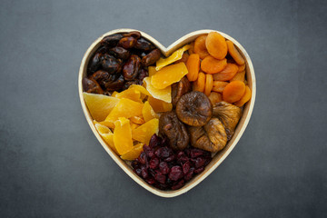 Dried fruits mix, in wooden heart shape box isolated on dark background. Top view of various dried fruits figs, apricots, mango, cranberries.