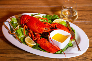 Boiled lobster served on a white ceramic platter with lemon slices and romaine lettuce, melted butter, and white wine in a stemless wine glass, against a wood background.