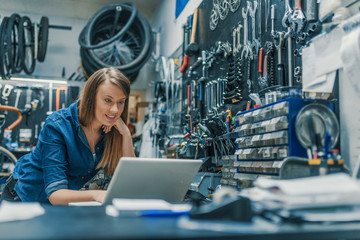 Portrait of beautiful working woman in the bicycle store. Young female mechanic with laptop. Female...