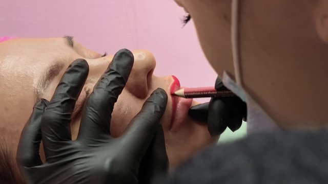 Preparation for permanent makeup. Draws a contour with a red pencil. Cosmetology procedure. Applying red permanent make up tattoo on young girl lips. Professional tattooist making permanent make up