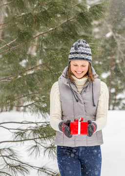 Beautiful, happy, smiling young woman holding Christmas gift present outdoors in snowy forest. She is wearing warm clothing a knit sweater, hat and mittens, denim jeans and an insulated vest.