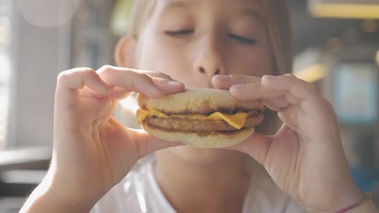 Beautiful happy hungry baby girl eating hamburger. Fast food concept. Tasty unhealthy burger sandwich in hands.