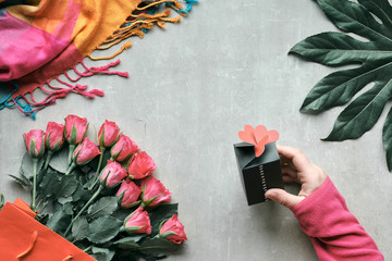 Flat lay, still life with bunch of rose flowers and exotic plant leaf. Hand holding small gift box with hearts on top. Top view on light stone background. Valentine, birthday or mother's day concept.
