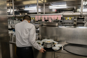 Waiter in the uniform is waiting for his orders. Big galley on a cruise ship. Black tray and a lot of plates around.