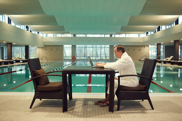 Full length side view of successful senior man using laptop while relaxing at luxury SPA resort with swimming pool in background, copy space