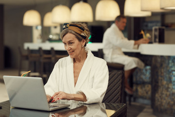 Portrait of modern senior woman smiling happily while using laptop in restaurant at luxury SPA resort, copy space