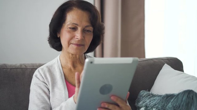 Mature caucasian woman sitting on sofa with electronic tablet in hand at home. Happy elderly female surfing web and texting in chat using silver digital device in apartment room. 60s brunnete is