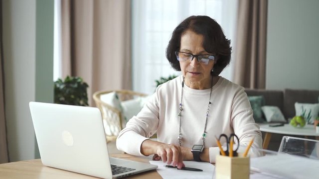 Mature american businesswoman using laptop sitting at table in home room. Portrait of elegant aged female working with computer, doing paperwork at desk in modern interior. 60s attorney having work