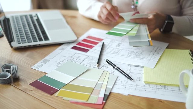 Mature woman expert working with color pantone sitting at table with laptop in room. Old female designer holding colour palette in hand, watching and choosing. New blueprint is at desk in modern