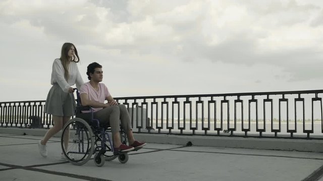 Sick disabled man is walking using wheelchair, woman is rolling it. They are resting together in park near river