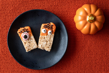 Creepy Halloween treat, two decorated crispy rice cookies on a black plate, orange ceramic pumpkin, orange background