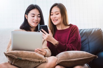 Young beautiful Asian women lesbian couple lover using smartphone and laptop shopping online together in living room at home with smiling face.Concept of LGBT sexuality with happy lifestyle together.