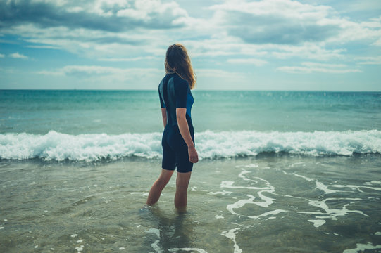 Young woman in wetsuit is standing on the beach with waves crashing in