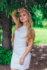 Fototapeta premium Portrait of a charming blond woman wearing beautiful white dress standing under fur tree.