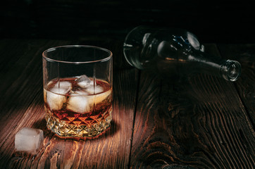 glass of whiskey with ice and an empty bottle on a wooden table
