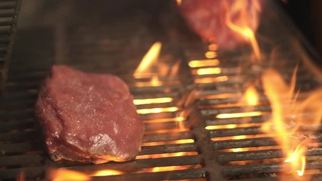 Two juicy pieces of raw meat are roasted on hot grill over an open flame. BBQ. Cook tongs turns Pork or beef cooked on grill. Barbecue outside house. Meat fillet is fried on fire on a cast iron grill.