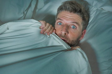  stressed and scared man alone in bed awake at night in fear after having a nightmare feeling paranoid holding the blanket in funny panic face expression