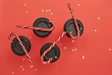 Paper coffee cup with sugar canes on a red background. Christmas background with Xmas candy canes and star confetti. Top view, flat lay, mockup. Copy space for text.