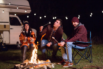 Man blowing a hot marshmallow while friends al laughing around camp fire