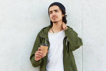 Fototapeta premium Photo of upset disappointed youngster looking aside, touching his neck with hand, feeling pain, being very tired, having lunchtime, spending leisure time alone, wearing casual items of clothes.
