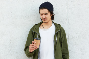 Fototapeta premium Portrait of confused unsatisfied hipster standing isolated over white wall outdoors, holding papercup with drink, undelighted with taste of drink, wearing casual clothes. Street style concept.