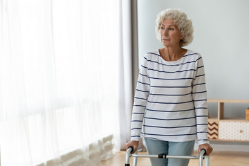 Thoughtful sad injured old woman standing alone using walking frame