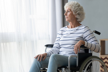 Sad paraplegic old woman sit on wheelchair look through window