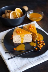Cheesecake with sea buckthorn berries and sea buckthorn sauce on a black plate with black teaspoons. In the background is a black bowl with a walnut and dried orange.